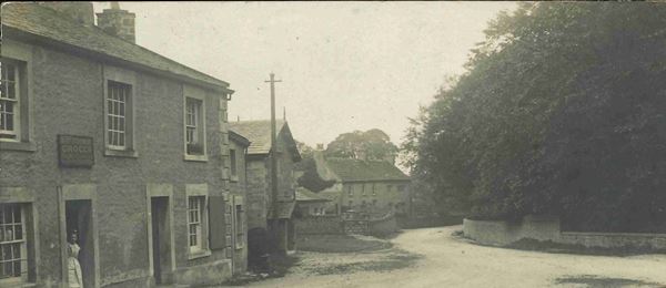 Black and white photo of the main road in Over Kellet in the 1920s. A woman stands in a shop doorway to the left. Large trees are surrounded by a stone wall to the right.